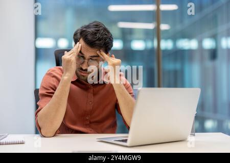 Geschäftsmann, der Stress und Erschöpfung am Arbeitsplatz erlebt und den Kopf mit beiden Händen hält. Die Büroumgebung umfasst ein Notebook, das Druck und Frustration im professionellen Umfeld widerspiegelt. Stockfoto