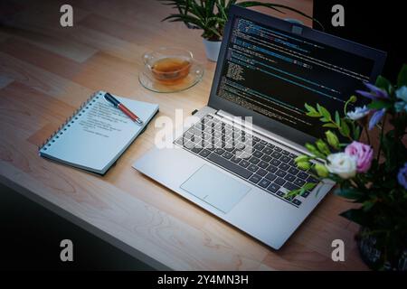Laptop mit Code auf dem Bildschirm und Notizblock mit handgeschriebenem Code auf einem Holztisch in der Nacht, Freiberufler Überstunden in der Software-Entwicklung oder Virenentwicklung Stockfoto