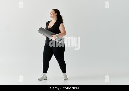 Eine elegante Frau in Übergröße steht selbstbewusst mit einer Yogamatte und lächelt in einem geräumigen Studio. Stockfoto