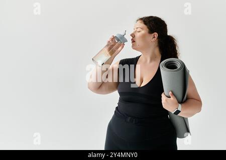 Eine selbstbewusste Frau in Übergröße genießt nach dem Training einen erfrischenden Drink, während sie eine Yogamatte hält. Stockfoto