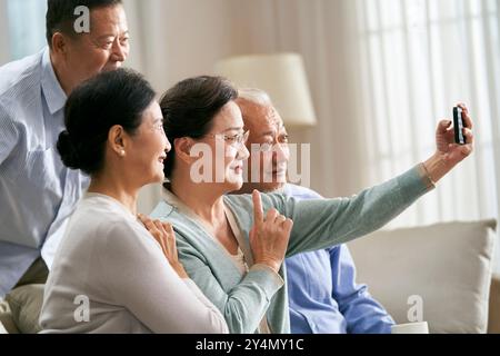 Gruppe glücklicher älterer asiatischer Leute zwei Paare, die zu Hause auf der Familiencouch sitzen und gemeinsam ein Selfie mit dem Handy machen Stockfoto