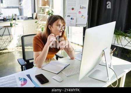 Der junge, nicht binäre Mensch beschäftigt sich mit einem Computer und denkt über Aufgaben in einem hellen Büro nach. Stockfoto