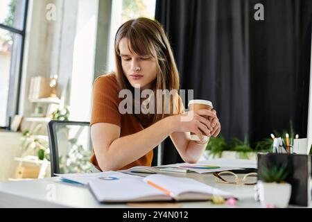 Eine junge, nicht binäre Person sitzt an einem Schreibtisch und schlürft Kaffee in einem lebendigen Arbeitsbereich. Stockfoto