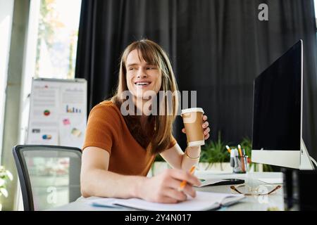 Mit einem warmen Lächeln schlürft ein nicht binärer Mitarbeiter Kaffee und nimmt an einem Schreibtisch Aufgaben wahr. Stockfoto