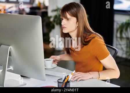 In einem modernen Büro arbeitet eine nicht binäre Person intensiv mit ihrem Computer, während sie Kaffee trinkt. Stockfoto