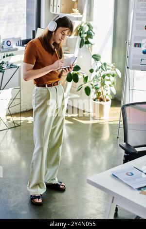 Eine junge, nicht binäre Person ist in einem modernen Büro mit Kopfhörern beschäftigt. Stockfoto