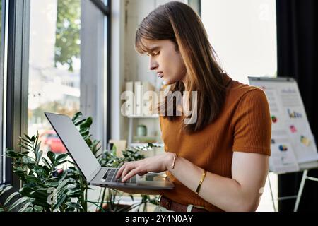 Ein fokussierter junger, nicht binärer Mensch greift in einem hellen und lebendigen Bürobereich mit seinem Laptop in Berührung. Stockfoto