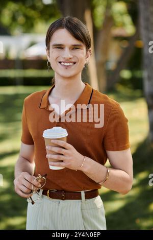 Eine junge, nicht binäre Person lächelt, während sie eine Kaffeetasse hält, umgeben von Grün. Stockfoto