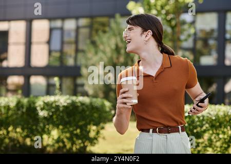 Eine Person lächelt hell, während sie Kaffee und Telefon in einem lebhaften Außenbüro hält. Stockfoto