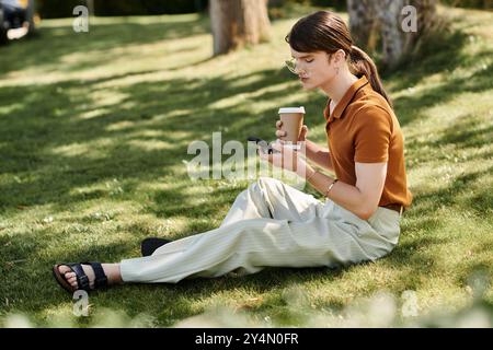 Auf dem Gras sitzend, schlürft eine nicht binäre Person Kaffee, während sie mit einem Smartphone in der Hand ist. Stockfoto