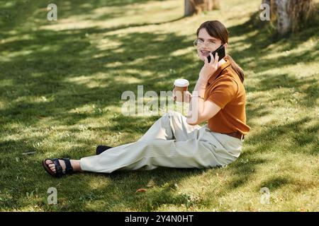 Eine junge, nicht binäre Person sitzt auf dem Gras und plaudert lebhaft am Telefon. Stockfoto