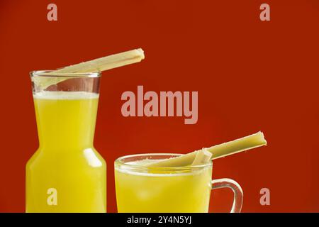 Nahaufnahme von zwei Flaschen Zuckerrohrsaft mit Zuckerrohrscheiben in einem Glas auf einem isolierten orangen Hintergrund Stockfoto