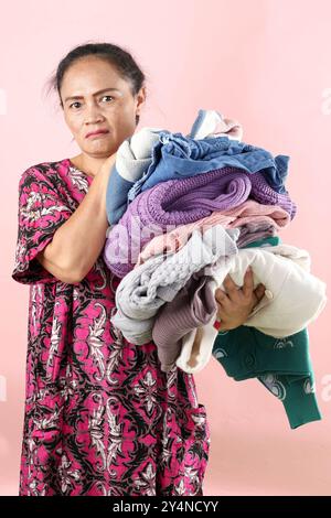 Asiatische Mutter Weearing Daster hält faltige Kleidung Wäsche Wäsche mit wütendem Gesichtsausdruck, auf rosa Hintergrund Stockfoto