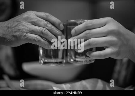 Zwei Hände, die zu Hause eine Gläser mit einem Schuss Whiskey anschlagen. Hände eines alten Mannes und eines jungen Mannes zünden Wodka an. Schwarzweißfoto. Zwei Gläser Stockfoto