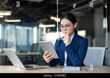 Asiatische Geschäftsfrau in blauem Anzug konzentriert sich auf die Verwendung eines Tablets in modernen Büros. Das professionelle Setting umfasst Laptop und Notizblock, in dem digitale Technologie für geschäftliche Aufgaben präsentiert wird. Stockfoto