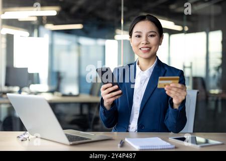 Selbstbewusste asiatische Geschäftsfrau führt Online-Zahlungen per Smartphone und Kreditkarte im Büro durch. Zeigt professionelle Einstellung beim Multitasking mit Laptop und Telefon Stockfoto