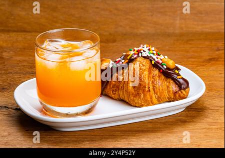 Blick auf kalten Orangensaft mit Eiswürfel in transparentem Glas mit köstlichem Croissant garniert mit Schokolade und Regenbogenstreuseln in Weißglas Stockfoto