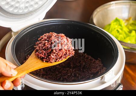 Blick aus einem hohen Winkel auf heiße, gedämpfte Reisbeere, Reisbeere in Holzlöffel vom geöffneten Reiskocher mit grünem Gemüse auf Holztisch. Stockfoto