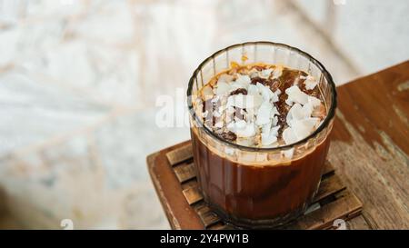 Kokos Eiskaffee in einem hohen Glas mit Kokosnusswürfel und Kaffeebohnen auf einem Holztisch. Stockfoto