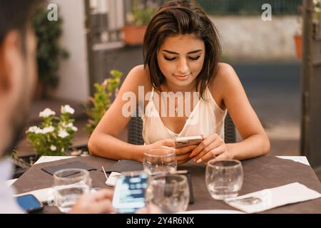 Attraktive junge Frau in lässiger Sommerkleidung mit Smartphone an einem Kaffeetisch im Freien. Moderne Lifestyle-Szene, die die Nutzung mobiler Technologien zeigt, soc Stockfoto