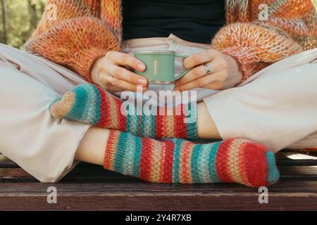 Frau mit Kaffeetasse, die in gestrickter Kleidung auf der Bank sitzt, Blick von oben. Mädchen mit Kaffee und überkreuzten Beinen. Person in gestreiften Stricksocken und Strickjacke Stockfoto