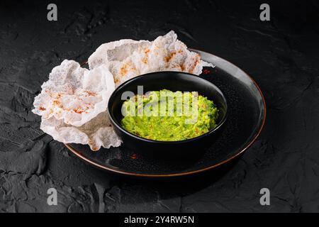 Leuchtend grüne Guacamole in einer schwarzen Schüssel neben knusprigen Reischips auf dunklem, strukturiertem Hintergrund Stockfoto