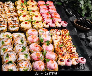Farbenfrohe Auswahl an Sushi und Brötchen, wunderschön präsentiert auf einem Schiefertablett inmitten grüner Blätter Stockfoto