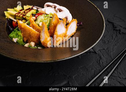 Exquisiter gebratener Hähnchensalat mit frischem Gemüse und Avocado, serviert auf einem dunklen Teller mit stilvoller Präsentation Stockfoto