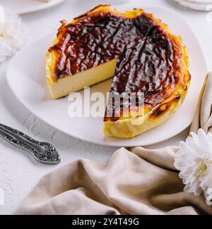 Verbrannter Gourmet-Käsekuchen aus baskenland mit einer Scheibe auf weißem Teller, begleitet von zündeten Kerzen und Blumen Stockfoto