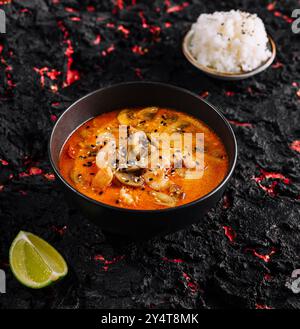 Lebendige Schüssel mit scharfer thai-Suppe mit Reis, garniert mit Samen und Limette, präsentiert auf einer dunklen, strukturierten Oberfläche Stockfoto