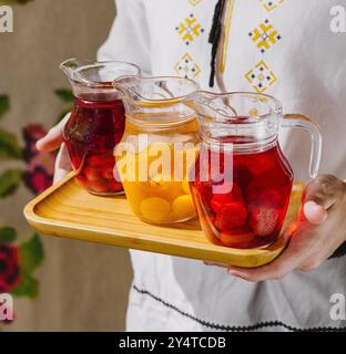 Die Person stellt drei Krüge mit erfrischendem, mit Obst angereichertem Wasser auf einem Holztablett aus nächster Nähe vor Stockfoto