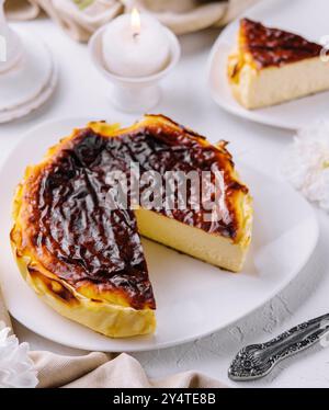 Verbrannter Gourmet-Käsekuchen aus baskenland mit einer Scheibe auf weißem Teller, begleitet von zündeten Kerzen und Blumen Stockfoto