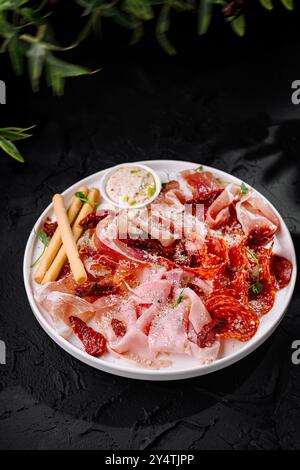 Gourmet-Auswahl an Räucherfleisch, serviert mit Brotstangen und Sauce vor dunkler Kulisse Stockfoto