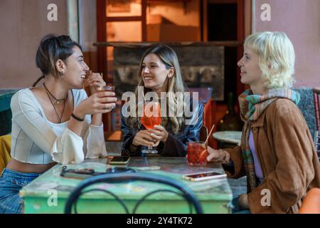 Drei verschiedene junge Freundinnen lachen und genießen Cocktails in einem trendigen Café. Multiethnische Gruppe mit Freundschaft, sozialer Interaktion und m Stockfoto