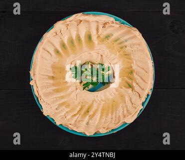 Cremiger Hummus mit Olivenöl und Petersilienblättern serviert auf blauem Teller auf schwarzem Holztisch Stockfoto