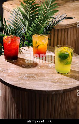 Drei bunte Cocktails sitzen auf einem Travertintisch mit tropischen Pflanzen im Hintergrund Stockfoto