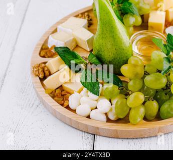 Auf einem weißen Holztisch befindet sich eine wunderschöne Wurstplatte mit verschiedenen Käsesorten, frischen Trauben, einer Birne und Honig Stockfoto