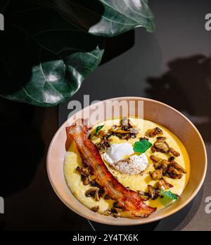 Köstliche Polenta mit gebratenen Pilzen, knusprigem Speck und pochiertem Ei, gewürzt mit frischen Kräutern, serviert auf einem Tisch Stockfoto