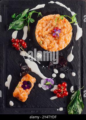 Lachs-Tartar wird auf einem schwarzen Schieferteller serviert, der mit Kräutern, Blütenblättern und Johannisbeere dekoriert ist Stockfoto