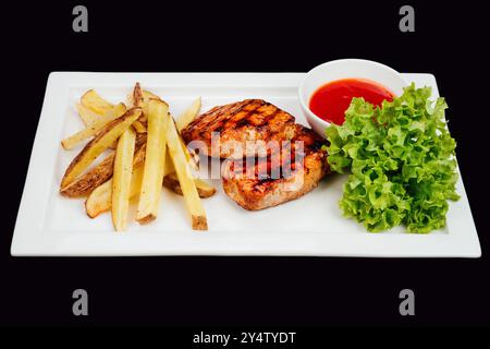 Gegrillte Schweinefleisch-Steaks werden mit Pommes frites, Salat und Tomatensauce auf weißem, rechteckigem Teller auf schwarzem Hintergrund serviert Stockfoto