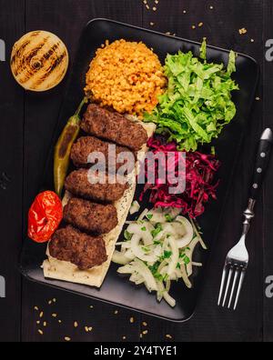 Traditionelle türkische Kofta-Kebabs, serviert mit frischem Gemüse, Pita-Brot und gewürztem Bulgur auf einem schwarzen Teller Stockfoto