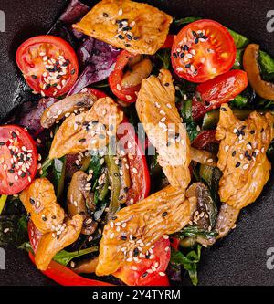 Salat mit warmem Huhn, Gurken, Tomaten, Pilzen, Kräutern und Sesamsamen wird auf einem schwarzen Teller serviert Stockfoto