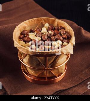 Kupferkorb, der mit gerösteten Erdnüssen überfüllt ist und auf einer braunen Serviette auf einer dunklen Holzoberfläche sitzt Stockfoto
