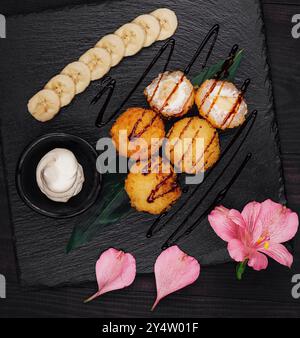 Frittierte Bananen- und Frischkäse-Sushi-Bällchen werden auf einer schwarzen Steinplatte mit Schokoladensirup und in Scheiben geschnittenen Bananen serviert Stockfoto