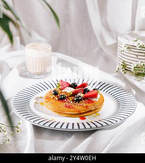 Pfannkuchen mit Schlagsahne, Beeren und Pistazien stehen auf weißer Tischdecke in der Nähe von Cappuccino und Palmblättern Stockfoto