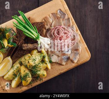 Holzbrett mit gesalzenem Hering, gekochten Kartoffeln, Roggenbrot, eingelegten Gurken, frischen Zwiebeln und Zitronen auf einem dunklen Holztisch Stockfoto