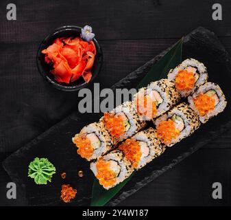 Sushi-Brötchen mit rotem Kaviar und Sesamsamen, serviert auf schwarzem Steinschiefer auf dunklem Holztisch mit Ingwer, Wasabi und weißer Orchidee Stockfoto