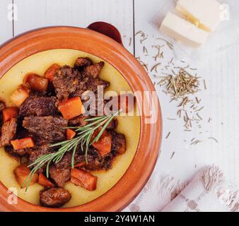 Köstlicher Rindereintopf mit Polenta und Gemüse in einer rustikalen Steingutform, garniert mit Rosmarin Stockfoto