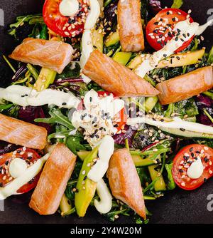 Köstlicher Salat mit gebratenem Thunfisch, Avocado und Kirschtomaten, bedeckt mit Sesamsamen und Dressing, serviert auf schwarzem Teller Stockfoto