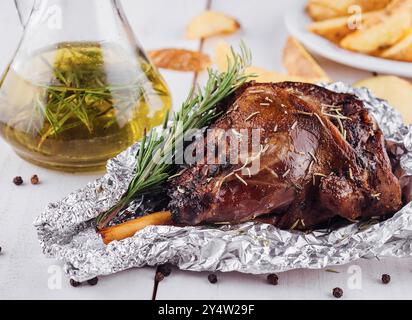 Köstliche gebratene Lammkeule mit Rosmarin und Gewürzen, serviert auf einem weißen Holztisch mit gebratenen Kartoffelkeimen Stockfoto